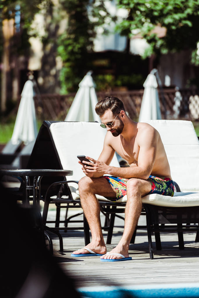 デッキチェアに座ってスマートフォンを使用してハンサムなひげの男の選択的な焦点  - 写真・画像