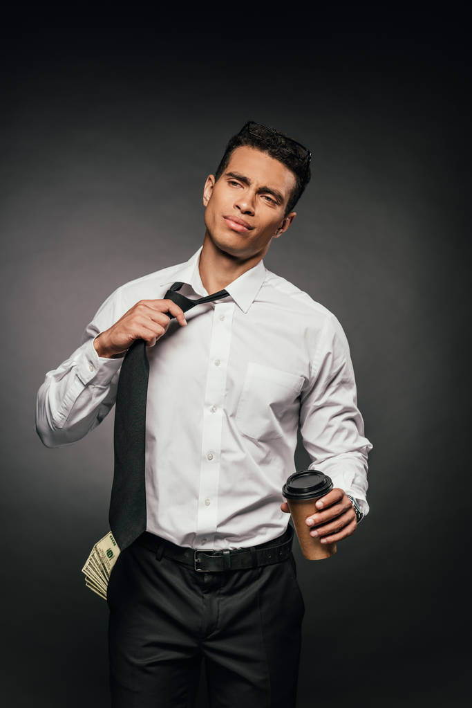 hombre de negocios afroamericano con dinero en efectivo en el bolsillo sosteniendo café para ir y desatar la corbata sobre fondo oscuro
 - Foto, imagen