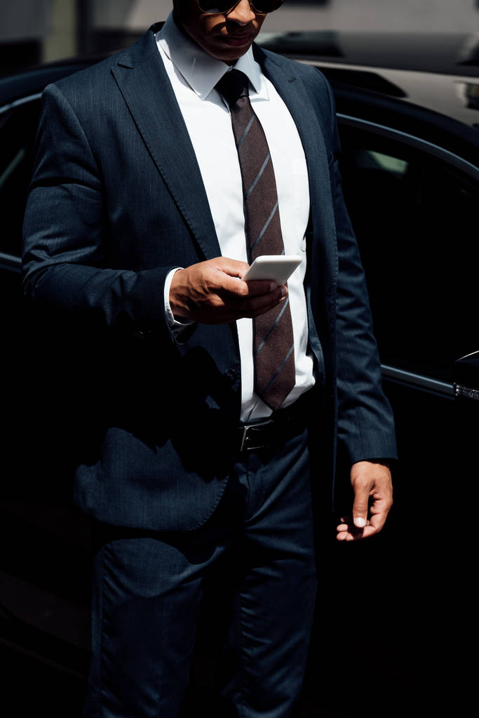 обрізаний вид афроамериканського бізнесмена в костюмі з використанням смартфона в сонячний день
 - Фото, зображення