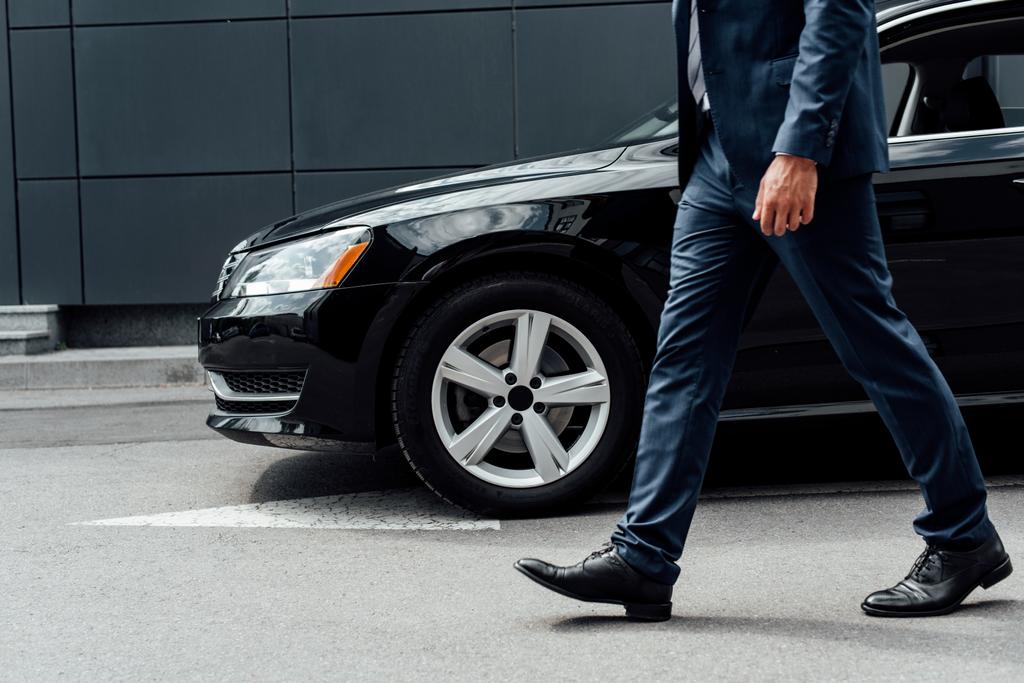 黒い車の近くの駐車場に沿って歩くスーツを着たアフリカ系アメリカ人ビジネスマンのトリミングされたビュー - 写真・画像