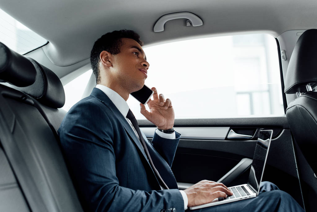 uomo d'affari afroamericano in giacca e cravatta utilizzando il computer portatile mentre parla su smartphone in auto
 - Foto, immagini