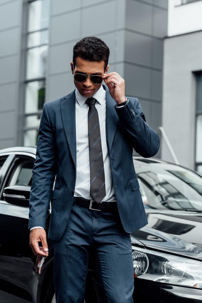 guapo hombre de negocios afroamericano en traje y gafas de sol caminando en el estacionamiento
 - Foto, Imagen