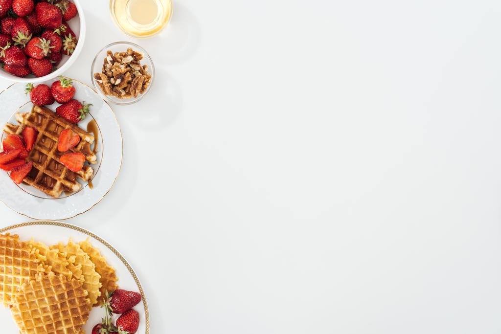 Blick von oben auf leckere Waffeln und Erdbeeren auf plattierten Schüsseln mit Honig und Nüssen auf weißen - Foto, Bild