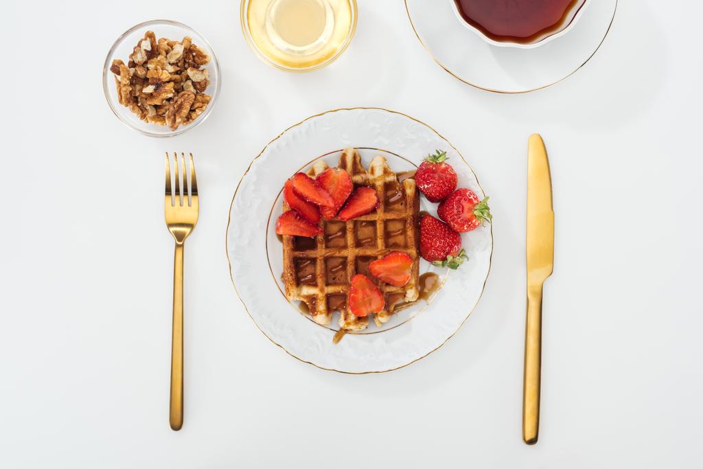 top view of served breakfast with waffle and strawberries on plate, honey and nits in bowls, cup of tea near fork and knife on white - Photo, Image