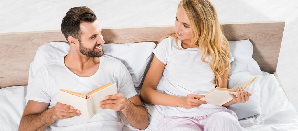 πανοραμικό πλάνο του πανέμορφης χαμογελαστή ζευγάρι που βρίσκεται στο κρεβάτι με τα βιβλία στο σπίτι  - Φωτογραφία, εικόνα