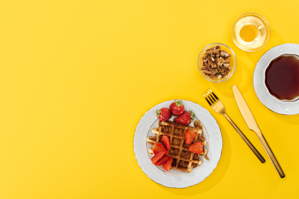 top view of waffle and strawberries, nuts, honey and tea near cutlery on yellow - Photo, Image