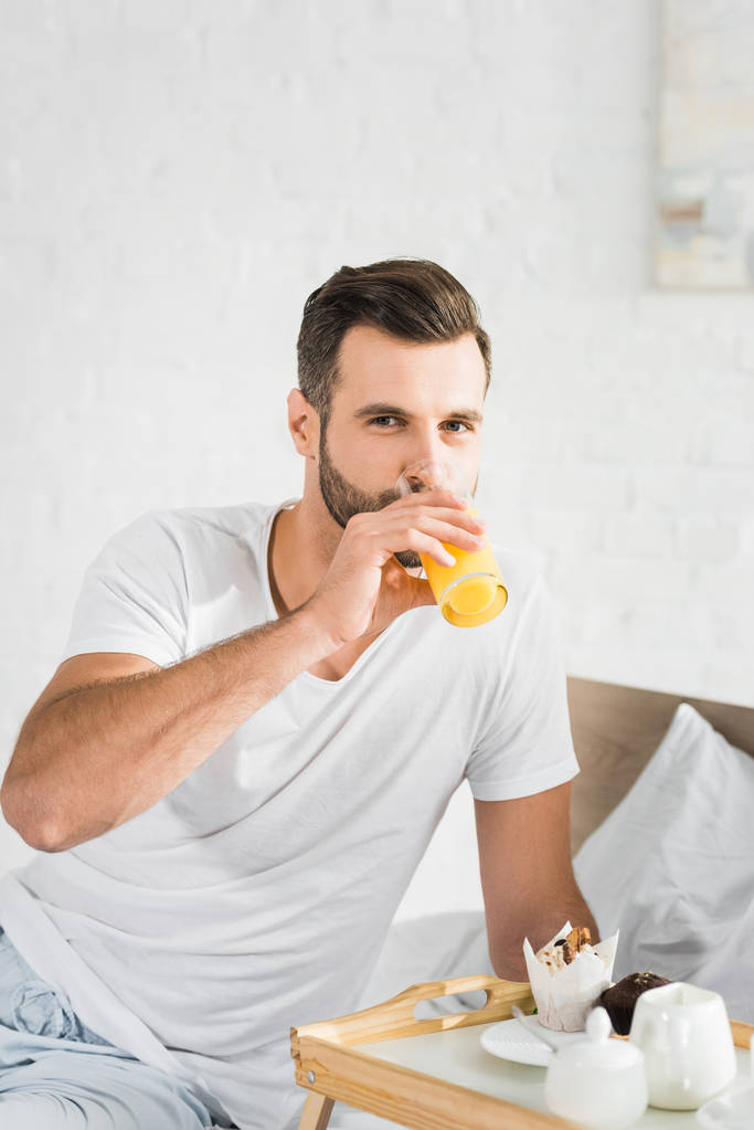 όμορφος άνθρωπος πίνοντας χυμό πορτοκάλι κοντά στο δίσκο τροφίμων κατά τη διάρκεια του πρωινού στο σπίτι - Φωτογραφία, εικόνα
