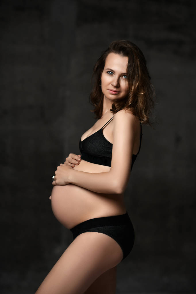 Beautiful young pregnant happy mother woman posing in underwear on dark  - Photo, Image