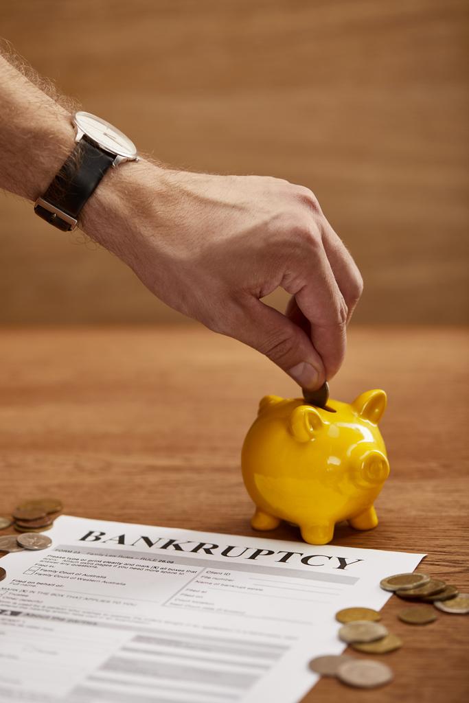 gedeeltelijke weergave van de man putting Coin in gele Piggy Bank in de buurt van faillissements formulier - Foto, afbeelding