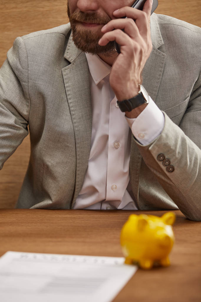 cropped view of businessman in suit talking on smartphone near yellow piggy bank - Photo, Image