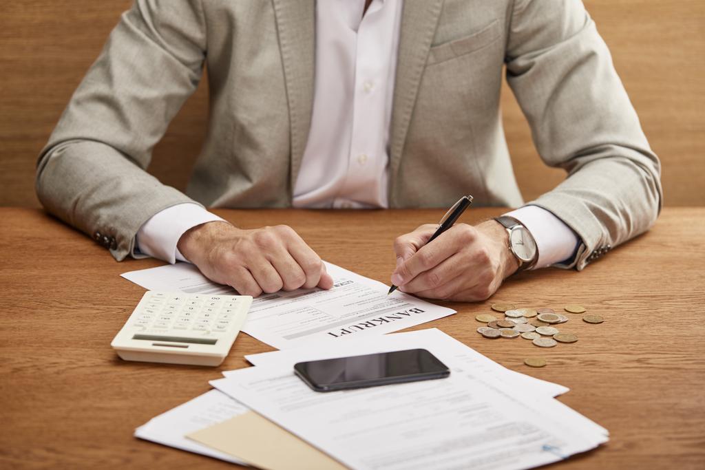 Ausgeschnittene Ansicht eines Geschäftsmannes im Anzug beim Ausfüllen des Konkursformulars am Holztisch - Foto, Bild