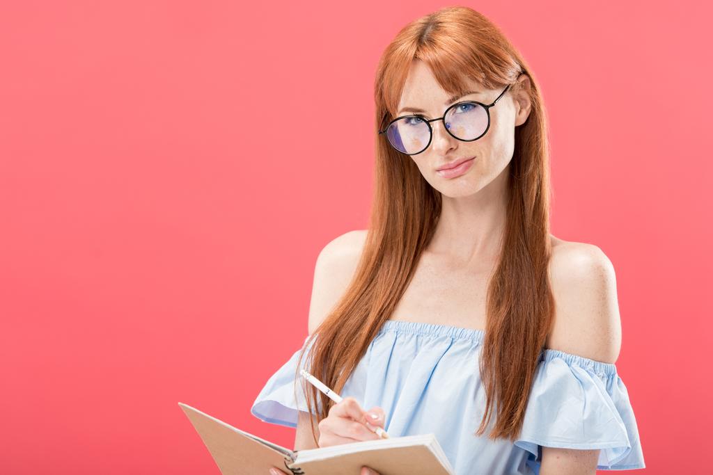 ελκυστική κοκκινομάλλα γυναίκα σε γυαλιά κρατώντας μολύβι και βιβλίο απομονωμένο σε ροζ - Φωτογραφία, εικόνα