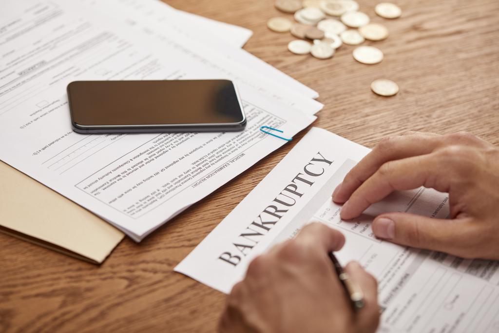 vista parcial del empresario llenando el formulario de bancarrota en la mesa de madera con teléfono inteligente y monedas
 - Foto, imagen