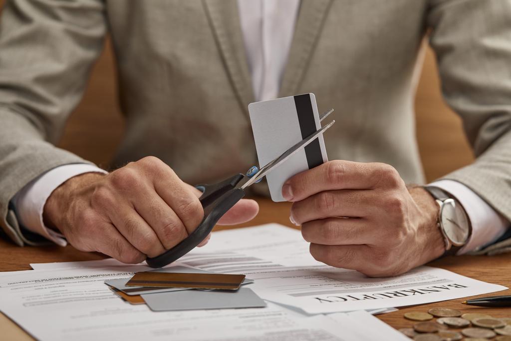 Teilansicht eines Geschäftsmannes im Anzug, der Kreditkarte mit der Schere an Holztisch schneidet  - Foto, Bild