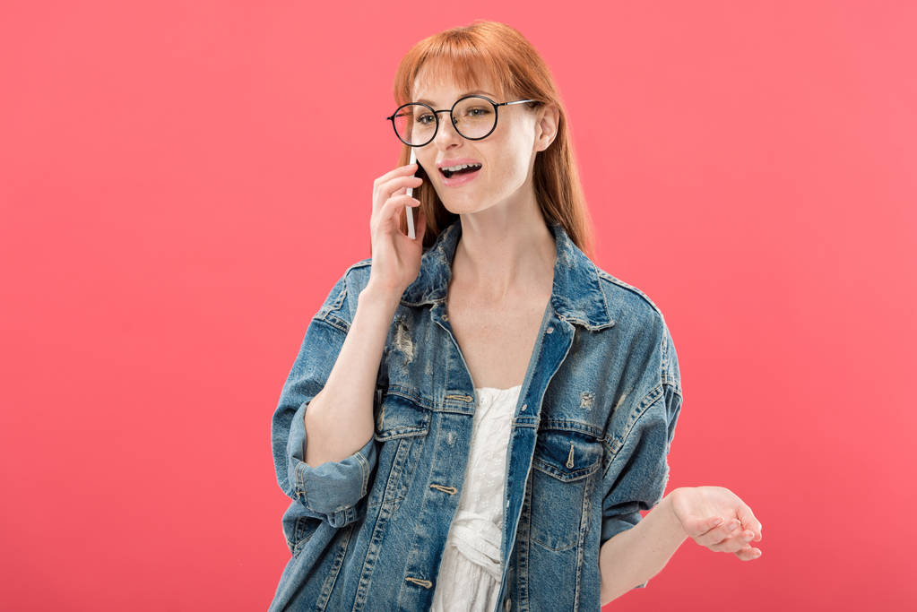 atractiva chica pelirroja en gafas y chaqueta de mezclilla hablando en el teléfono inteligente aislado en rosa
 - Foto, imagen