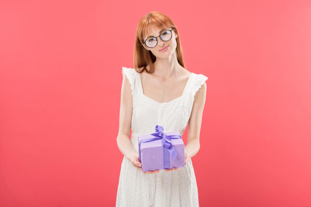 メガネと白いドレスで笑顔の赤毛の女の子の正面図誕生日プレゼントを保持し、ピンクに隔離されたカメラを見て - 写真・画像
