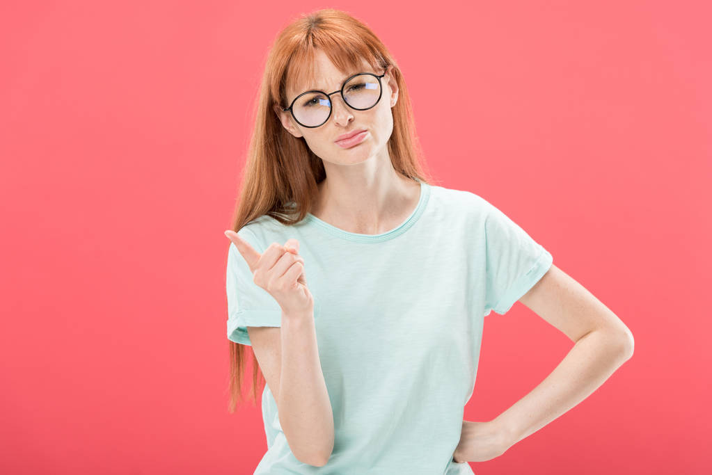 front view of serious redhead woman in glasses and t-shirt standing with hand on hip and shaking finger isolated on pink - Photo, Image