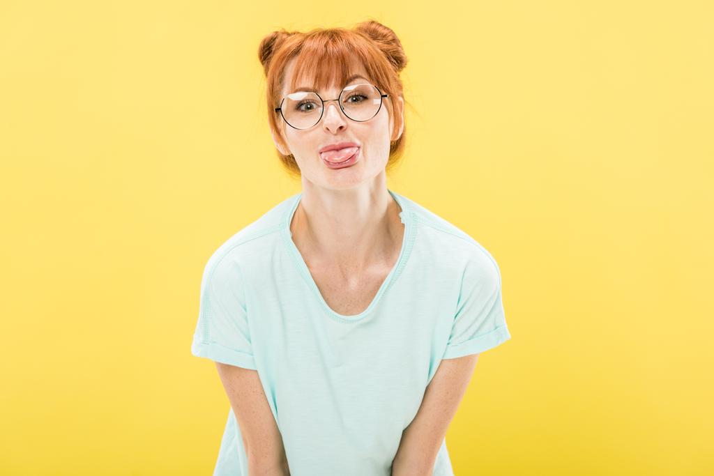 вид спереду весела руда дівчина в окулярах стирчить з язика і дивиться на камеру ізольовано на жовтому
 - Фото, зображення