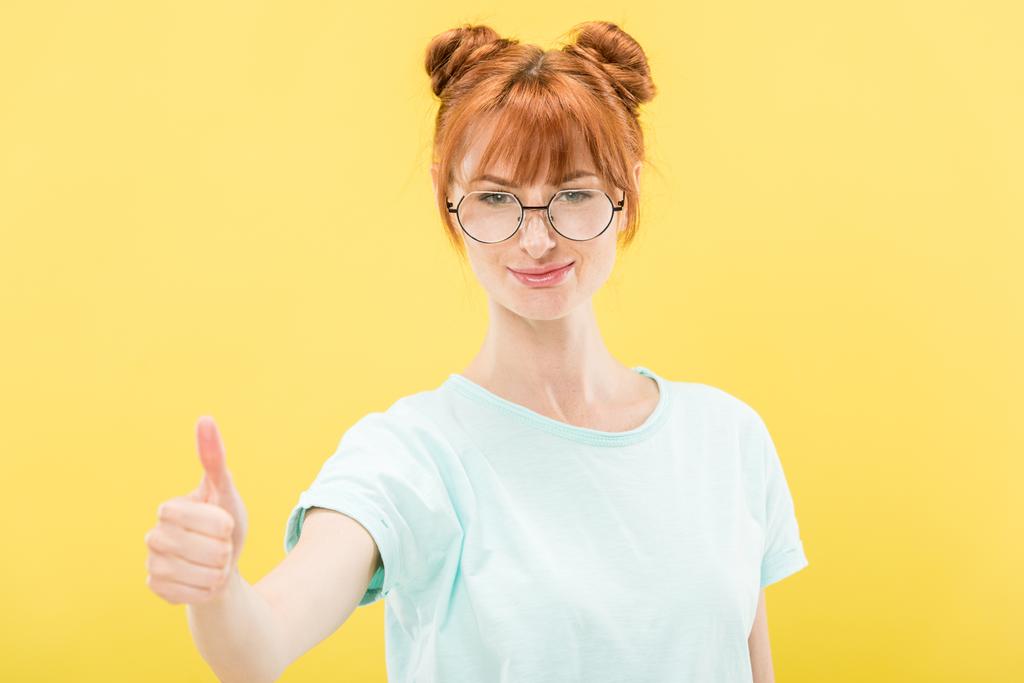 Vorderseite des lächelnden rothaarigen Mädchens in Brille und T-Shirt, das in die Kamera blickt und den Daumen vereinzelt auf gelb zeigt - Foto, Bild