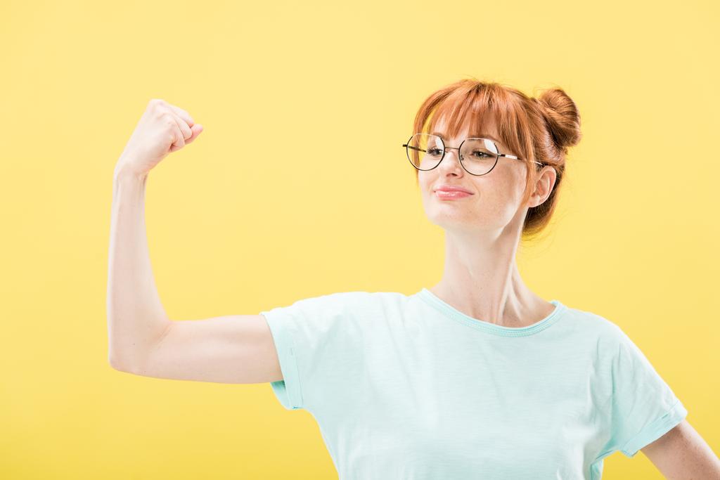 confident pleased redhead girl in glasses and t-shirt holding fist up isolated on yellow - Photo, Image