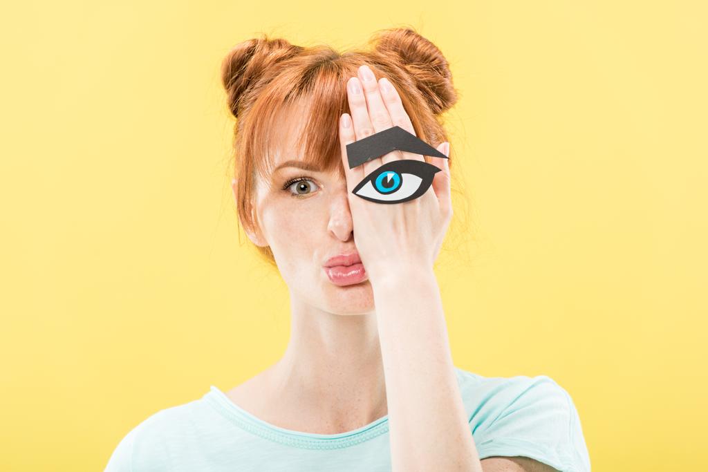 vista frontal de chica pelirroja sosteniendo ojo de papel y ceja y mirando a la cámara aislada en amarillo
 - Foto, Imagen