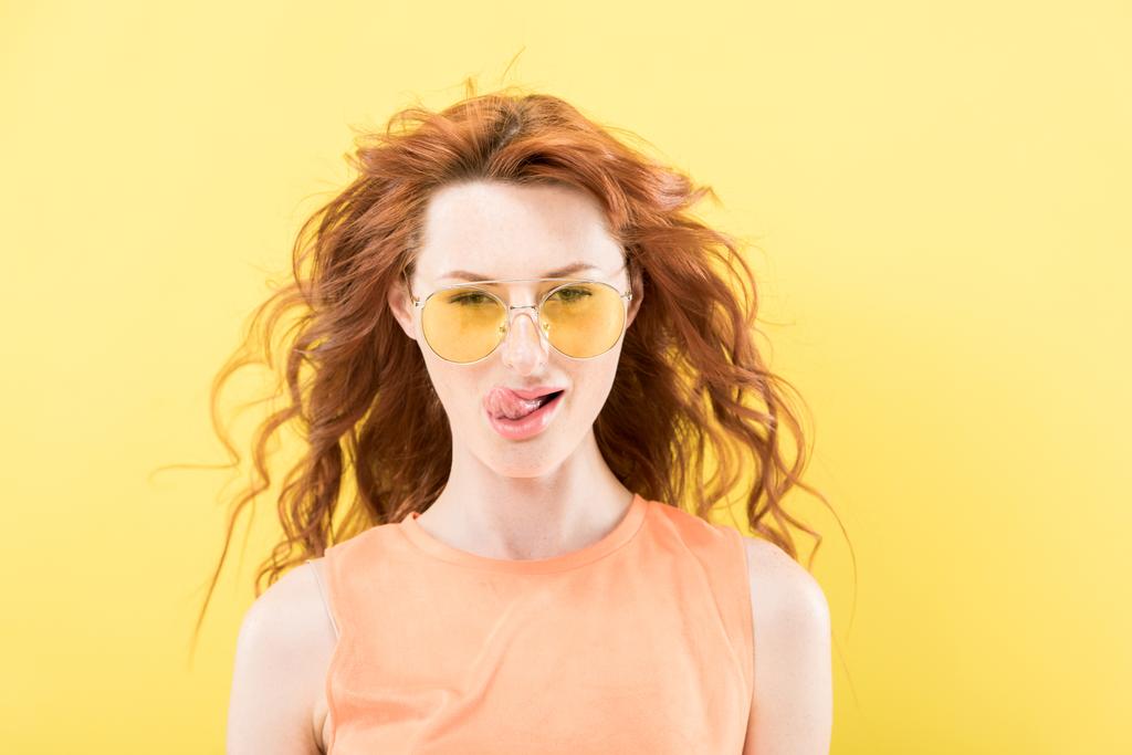 front view of curly redhead woman in sunglasses licking lips isolated on yellow - Photo, Image