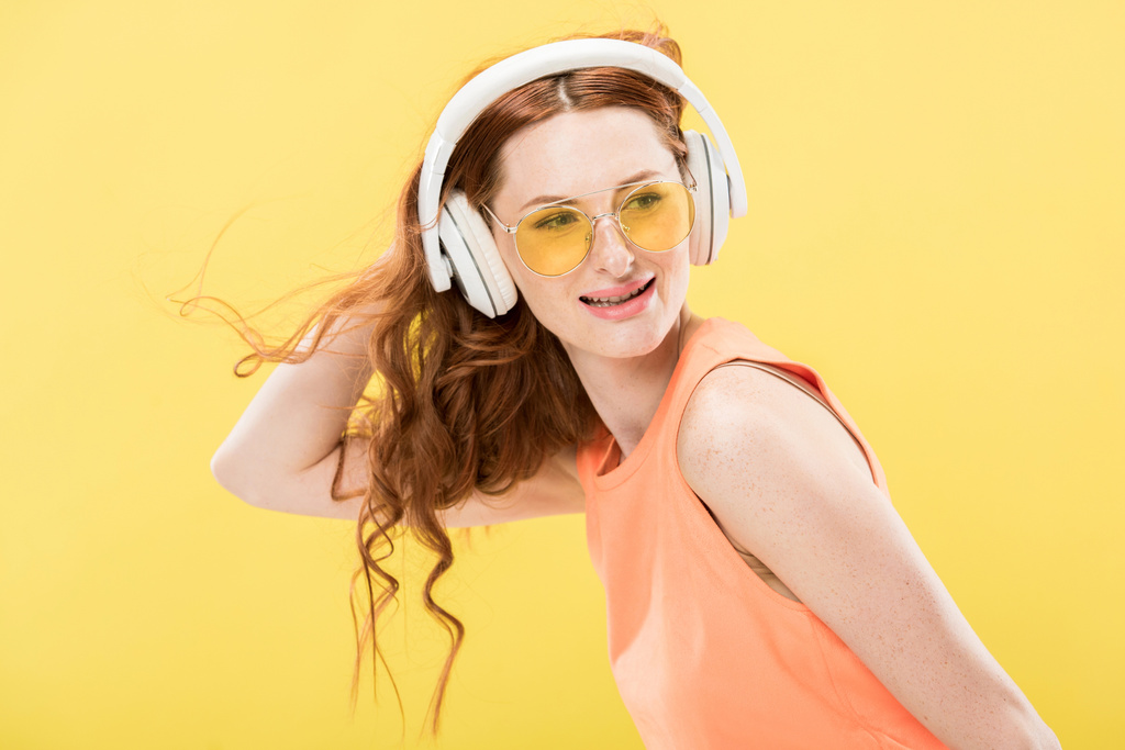 attraktive rothaarige Frau mit Sonnenbrille, die mit Kopfhörern Musik hört und isoliert auf gelb lächelt - Foto, Bild