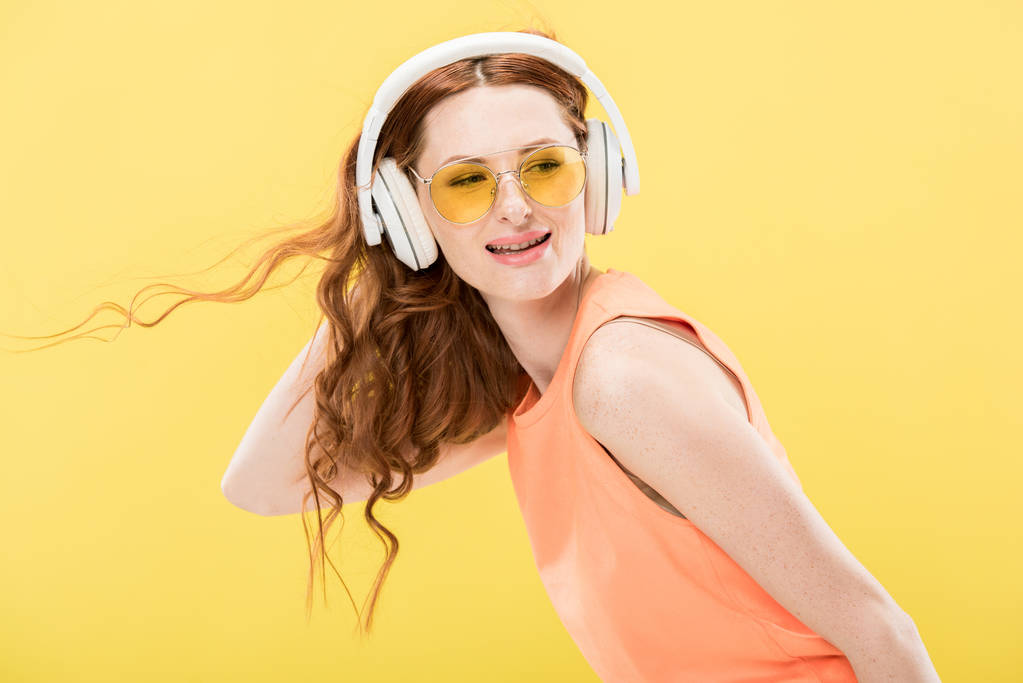 jolie rousse femme en lunettes de soleil écouter de la musique dans les écouteurs et sourire isolé sur jaune
 - Photo, image