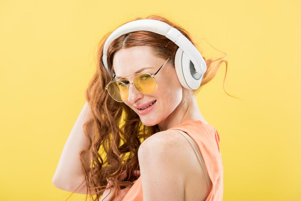 atractiva pelirroja en gafas de sol escuchando música en auriculares y sonriendo aislada en amarillo
 - Foto, Imagen