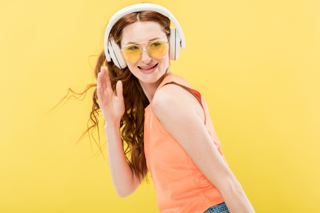 привлекательная рыжая женщина в солнечных очках слушает музыку в наушниках и улыбается изолированно на желтом
 - Фото, изображение