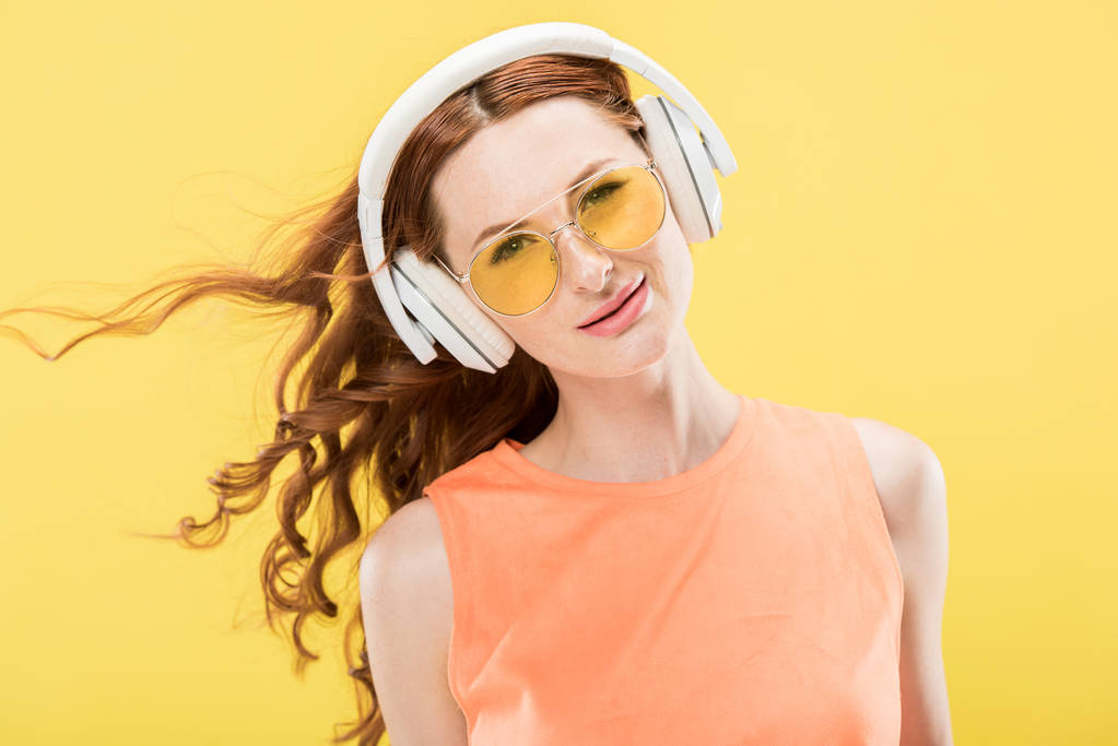 pohled na atraktivní zrzavou ženu v brýlích naslouchající hudbu ve sluchátkách a s úsměvem izolovaná na žluté - Fotografie, Obrázek