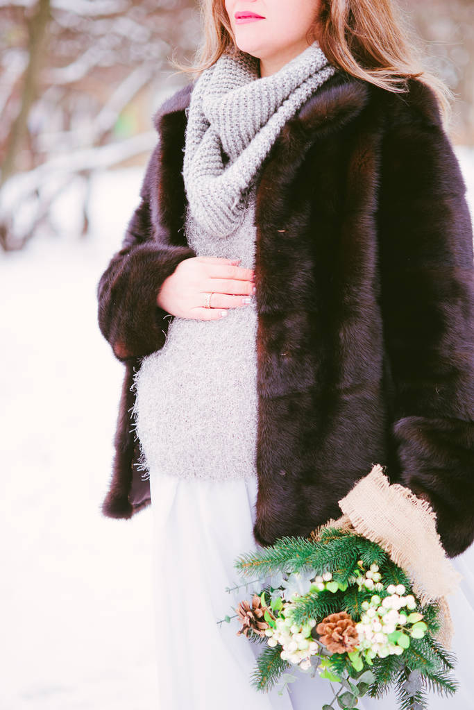 公園の雪の上で冬に花束を持つミンクのコートと長いドレスを着た若い妊娠中の女性 - 写真・画像