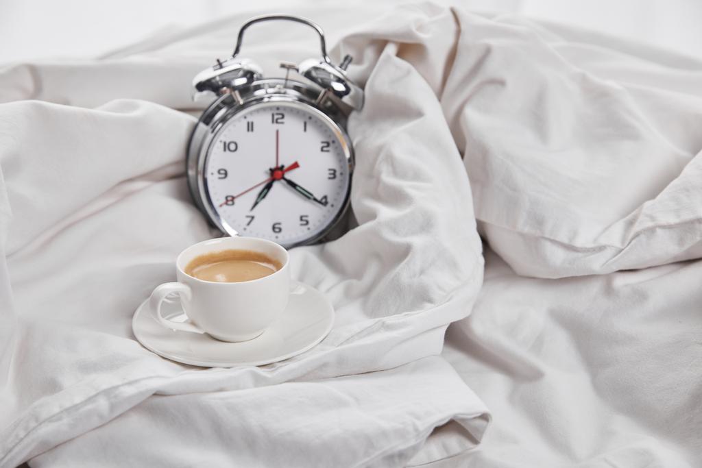καφέ σε λευκό Κύπελλο σε πιατάκι κοντά σε ασημένιο ξυπνητήρι σε λευκό κρεβάτι - Φωτογραφία, εικόνα