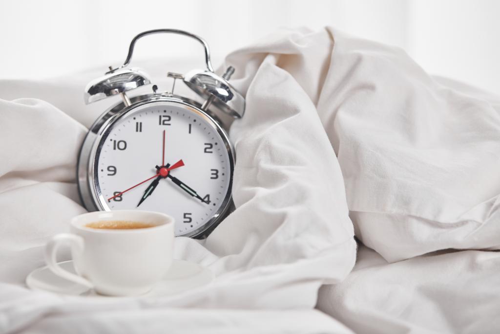 coffee in white cup on saucer near silver alarm clock in white blanket - Photo, Image