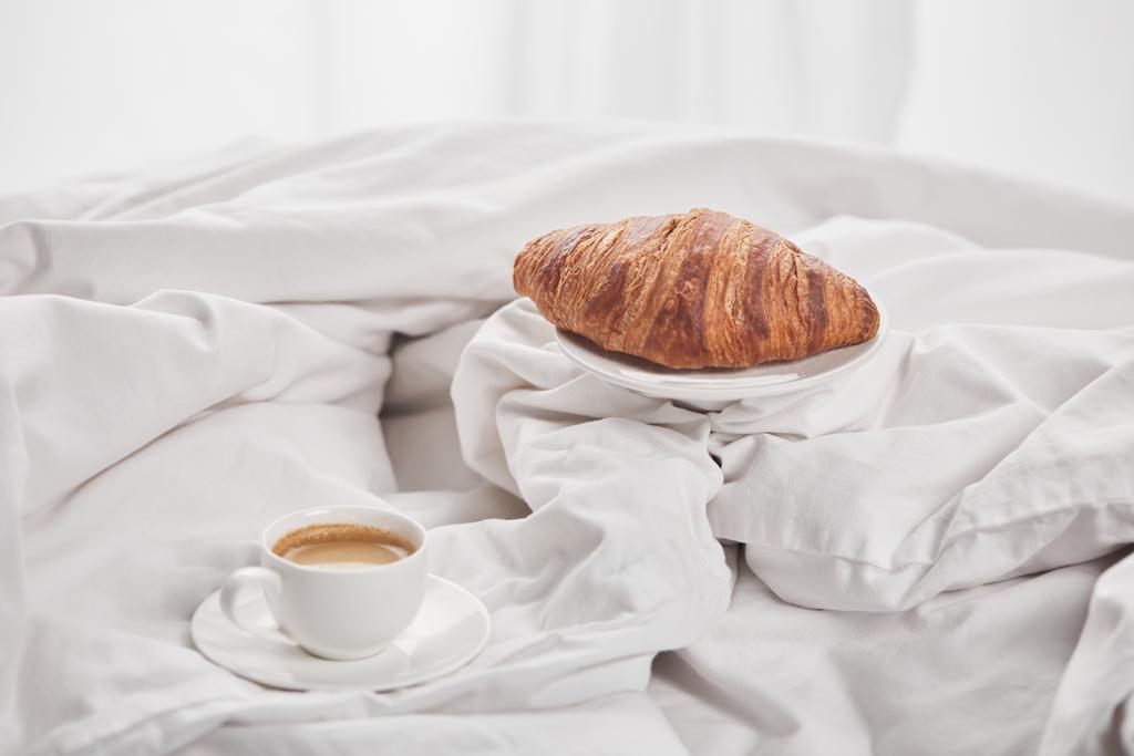 leckeres Croissant auf Teller neben Kaffee in weißer Tasse auf Untertasse im Bett - Foto, Bild