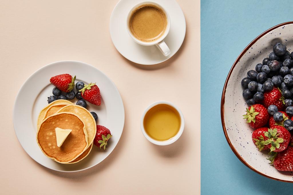 Pfannkuchen mit Beeren auf Teller, Schüssel mit Honig, Tasse Kaffee und Teller mit Beeren auf rosa und blauem Hintergrund  - Foto, Bild
