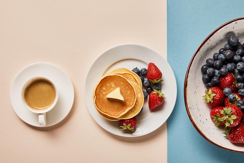 pohled na palačinky s bobule na desce blízko šálku kávy a talíř s jahodami a borůvkami - Fotografie, Obrázek
