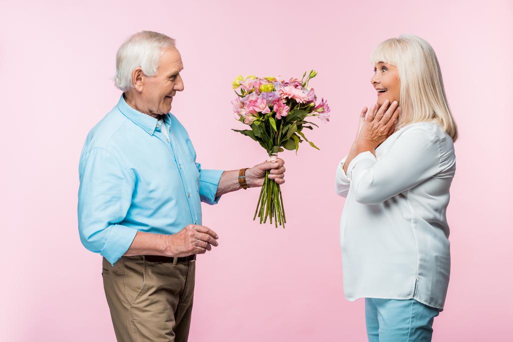 vrolijke Senior man geeft boeket met bloemen aan opgewonden vrouw op roze  - Foto, afbeelding