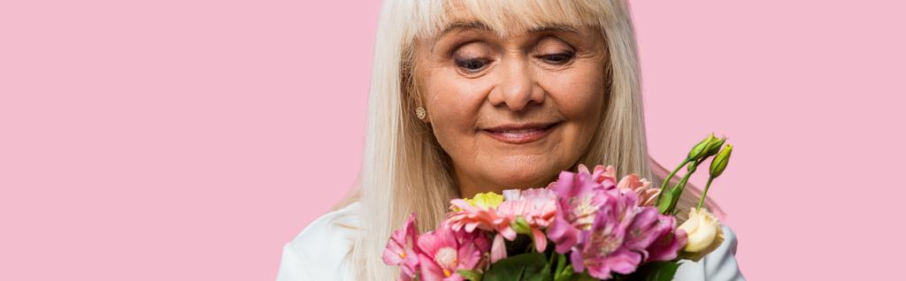 Panoramaaufnahme einer glücklichen Seniorin, die einen Strauß blühender Blumen betrachtet, isoliert auf rosa  - Foto, Bild