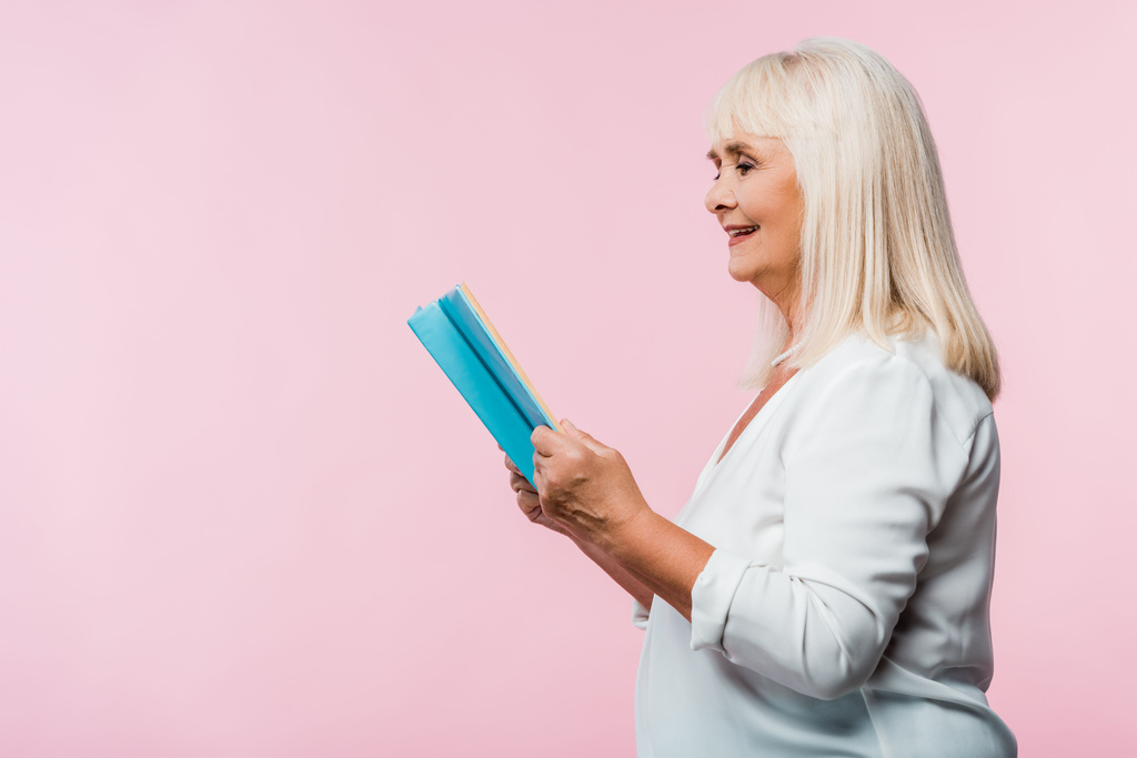 ピンクに隔離された灰色の髪の読書本を持つ陽気な引退した女性  - 写真・画像