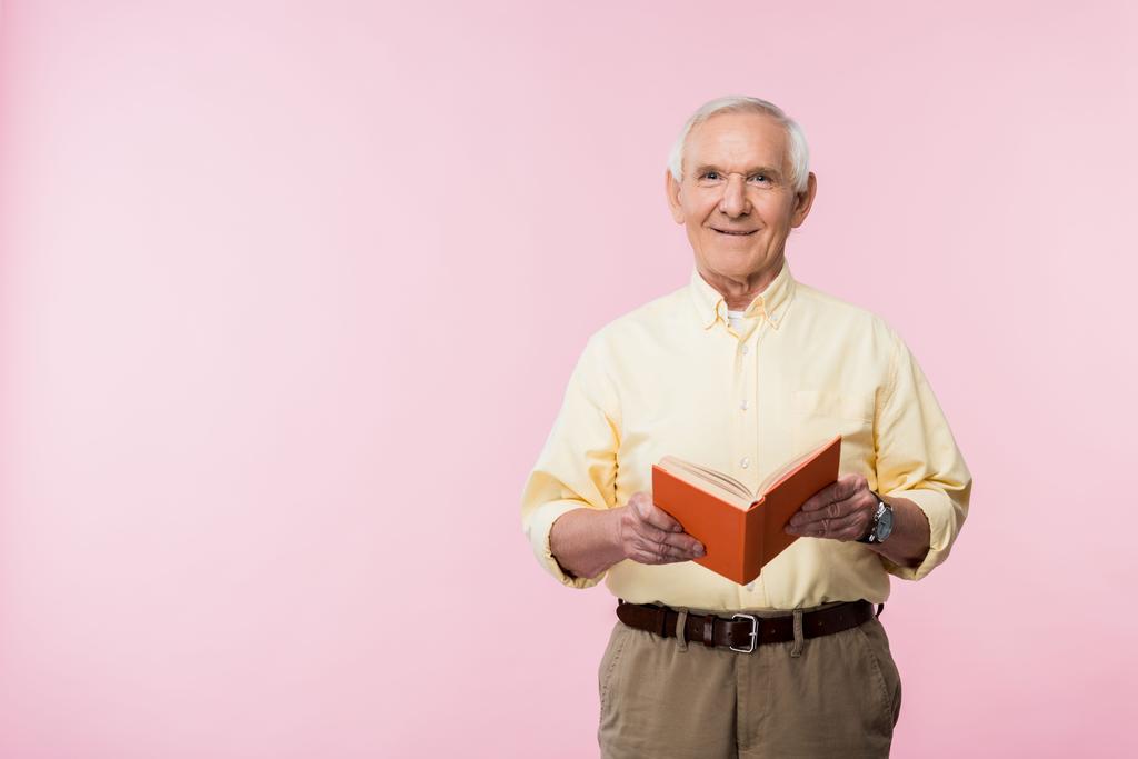 positieve gepensioneerde man Holding boek en glimlachend op roze  - Foto, afbeelding