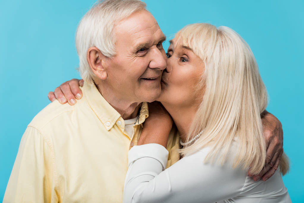 Starší žena líbala tvář veselého muže se šedými vlasy izolovanými na modrém  - Fotografie, Obrázek