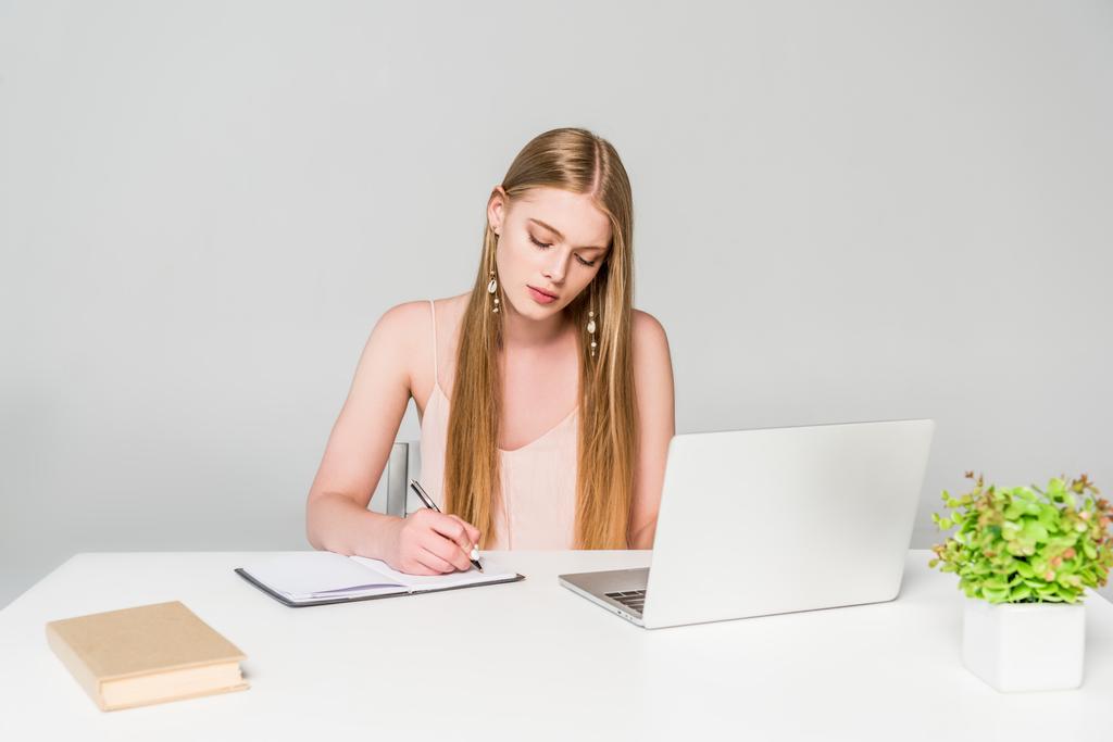 güzel kız bilgisayar masasında oturan ve gri üzerinde not defterine yazma - Fotoğraf, Görsel