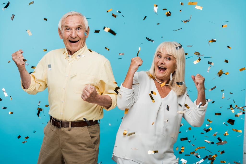glückliches Rentnerehepaar gestikuliert neben fallendem Konfetti auf blauem Grund  - Foto, Bild