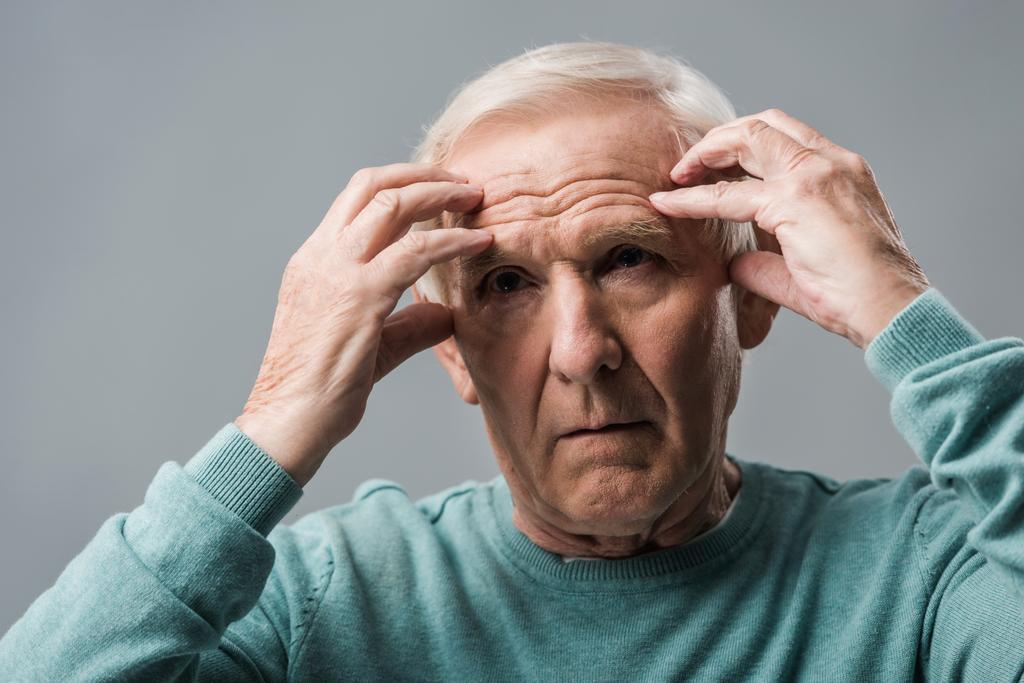 εξαντλημένο συνταξιούχος άνθρωπος κοιτάζοντας την κάμερα και αγγίζοντας το κεφάλι απομονωμένη σε γκρι  - Φωτογραφία, εικόνα