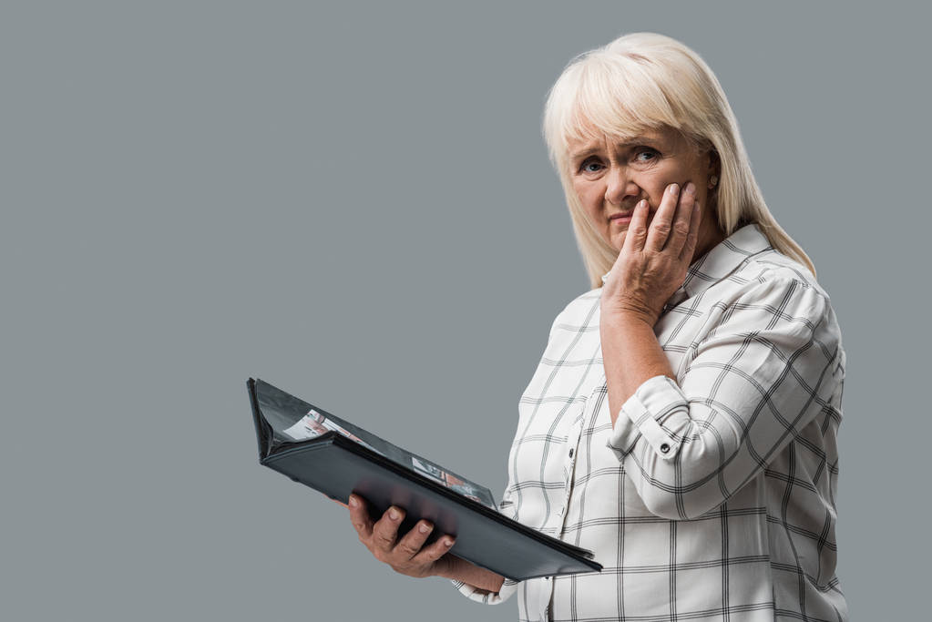 niezadowolona emerytowana kobieta trzyma album fotograficzny i dotykając twarzy na szaro  - Zdjęcie, obraz