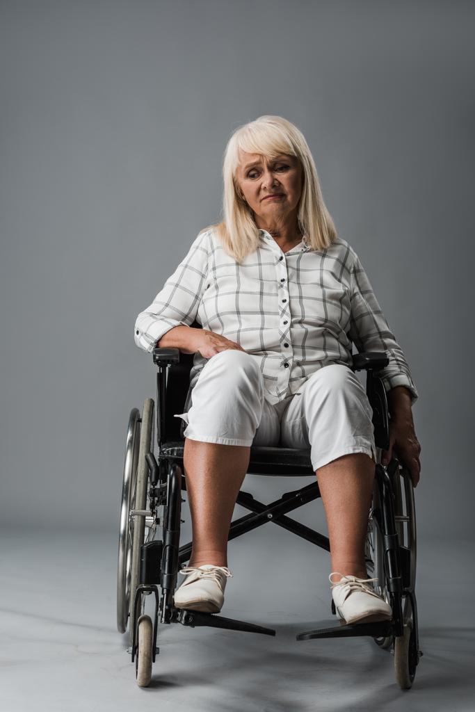 boos en gehandicapte gepensioneerde vrouw zittend in rolstoel op grijs  - Foto, afbeelding