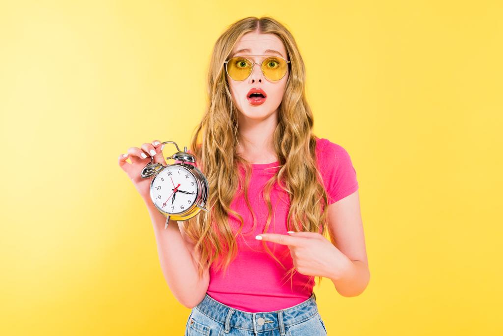 красива здивована дівчина в сонцезахисних окулярах, вказуючи пальцем на будильник На жовтому
  - Фото, зображення