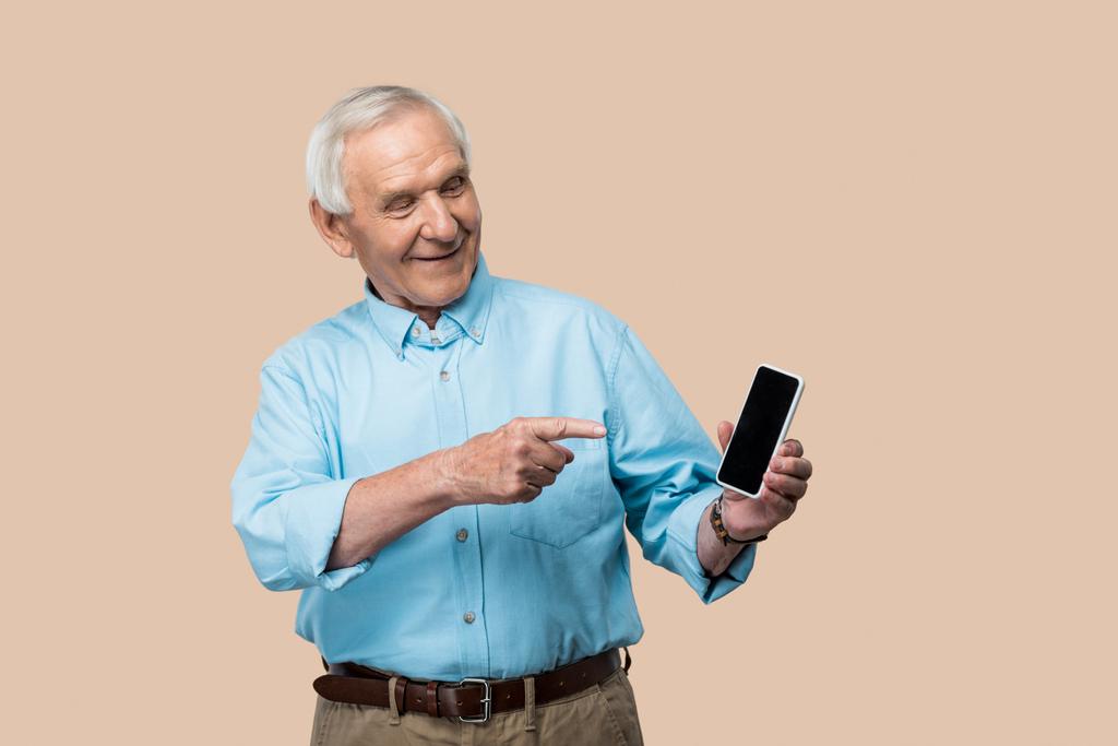 glücklicher Rentner mit grauen Haaren zeigt mit dem Finger auf Smartphone mit leerem Bildschirm isoliert auf beige  - Foto, Bild