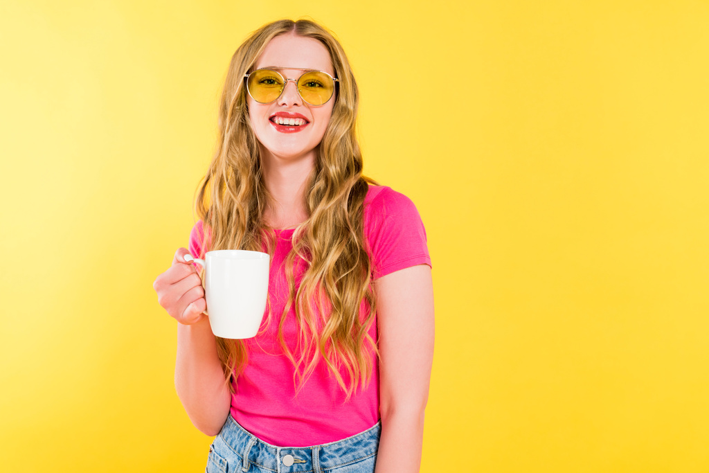 όμορφη χαμογελαστή κορίτσι σε γυαλιά ηλίου με φλιτζάνι καφέ απομονωμένη στο κίτρινο  - Φωτογραφία, εικόνα